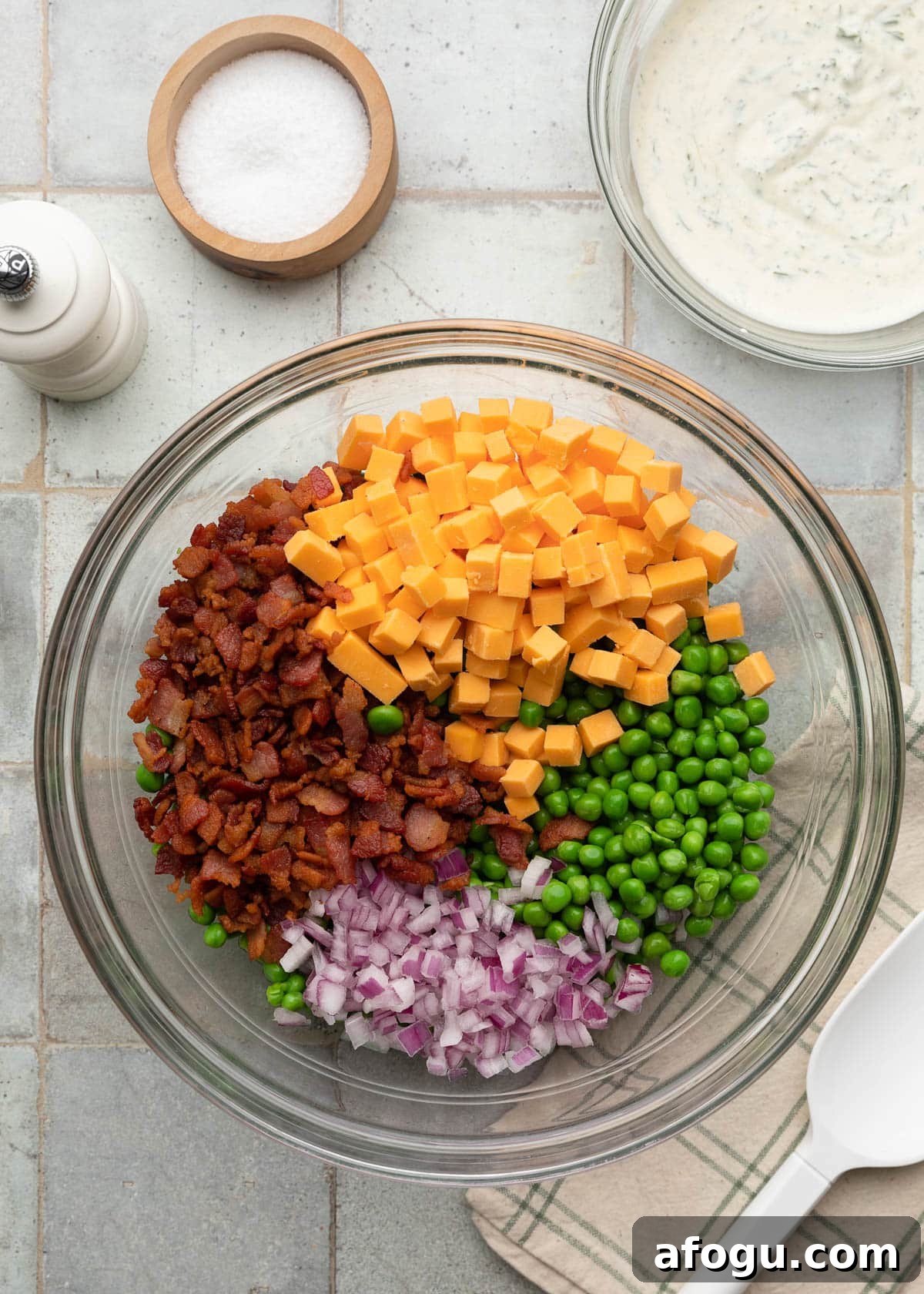 Top-down ingredient photo showing green peas, crispy chopped bacon, cheddar cheese cubes, and diced red onion arranged in a glass mixing bowl before tossing the salad together, with a separate bowl of creamy dill dressing beside it on the tiled surface.