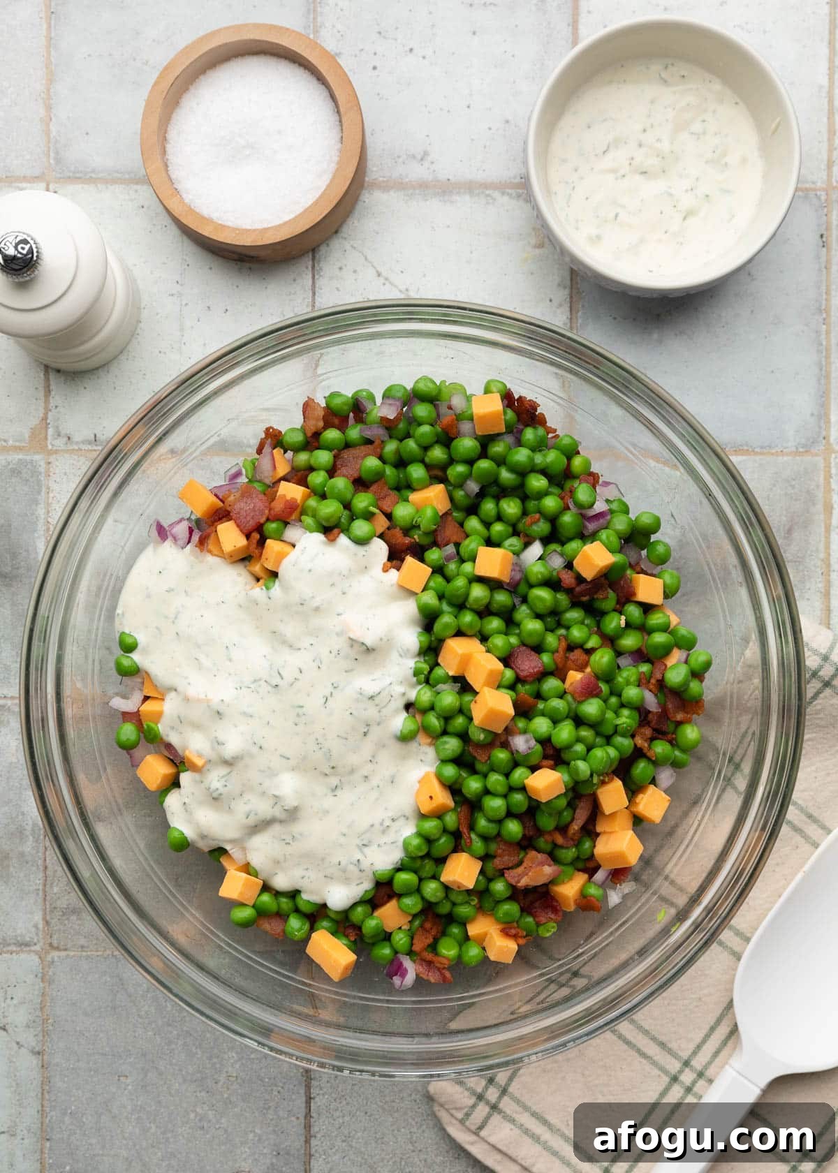 Top-down process photo of creamy dill dressing poured over a bowl of green peas, crispy bacon, cheddar cheese cubes, and diced red onion before mixing.