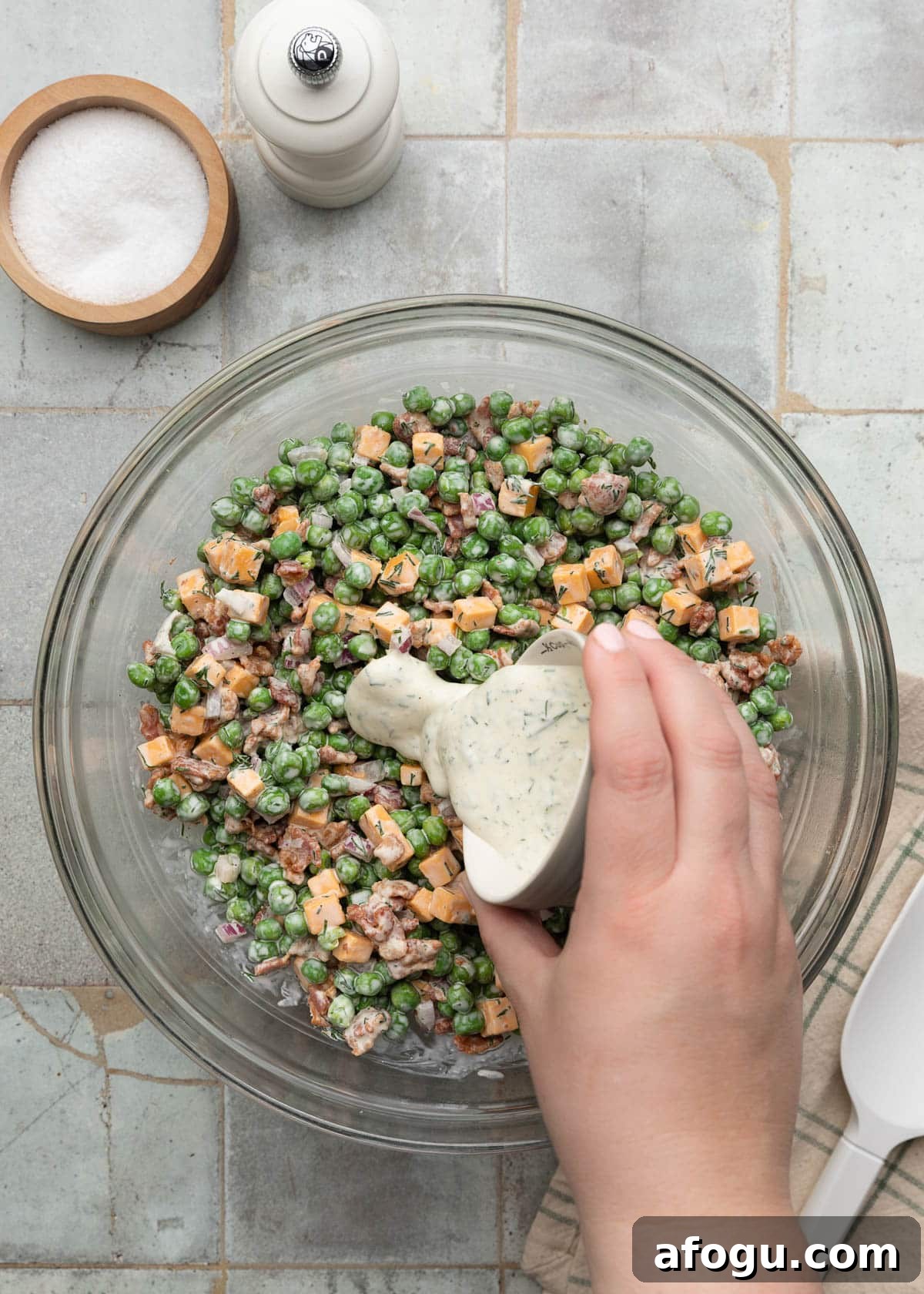 Top-down action photo showing creamy dill dressing being poured from a small bowl over a pea salad made with green peas, cheddar cheese cubes, crispy bacon, and diced red onion in a large glass mixing bowl.