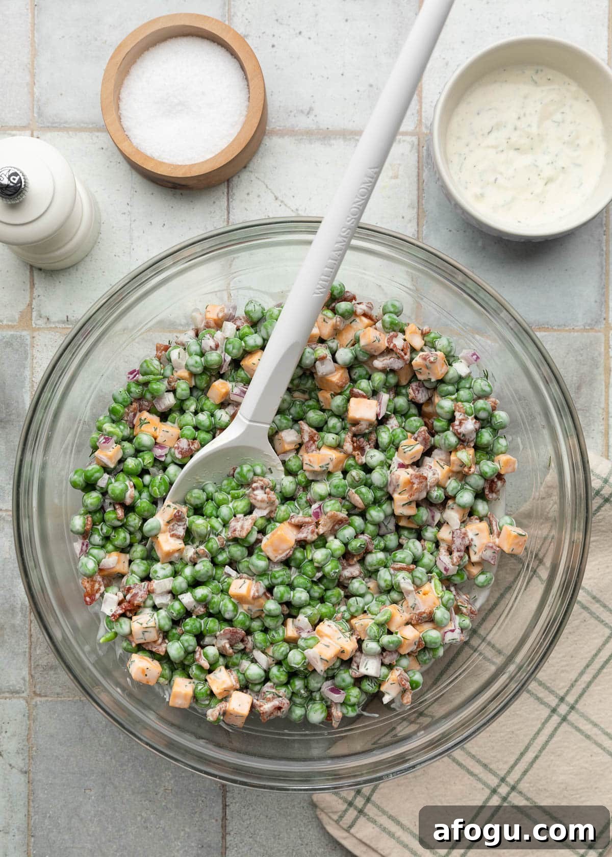 Top-down photo of creamy pea salad tossed in a glass mixing bowl with bright green peas, crispy bacon pieces, cheddar cheese cubes, diced red onion, and dill dressing, with a white rubber spatula resting in the bowl.