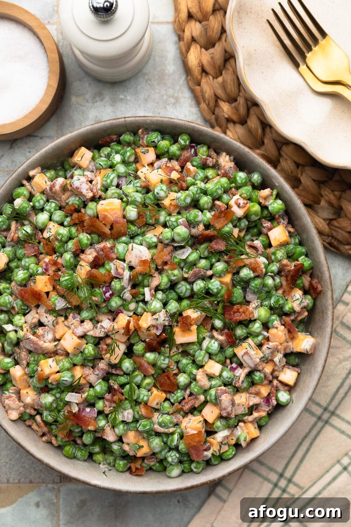 Top-down photo of creamy pea salad in a ceramic serving bowl, made with green peas, cheddar cheese cubes, crispy bacon, diced red onion, and fresh dill tossed in a creamy dressing. A white pepper mill, salt cellar, woven placemat, and gold fork are arranged around the bowl on a tiled surface.