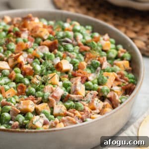 Close-up photo of creamy pea salad in a shallow ceramic serving bowl, made with green peas, cheddar cheese cubes, crispy bacon, diced red onion, and fresh dill in a creamy dressing. The salad is garnished with extra bacon and dill.