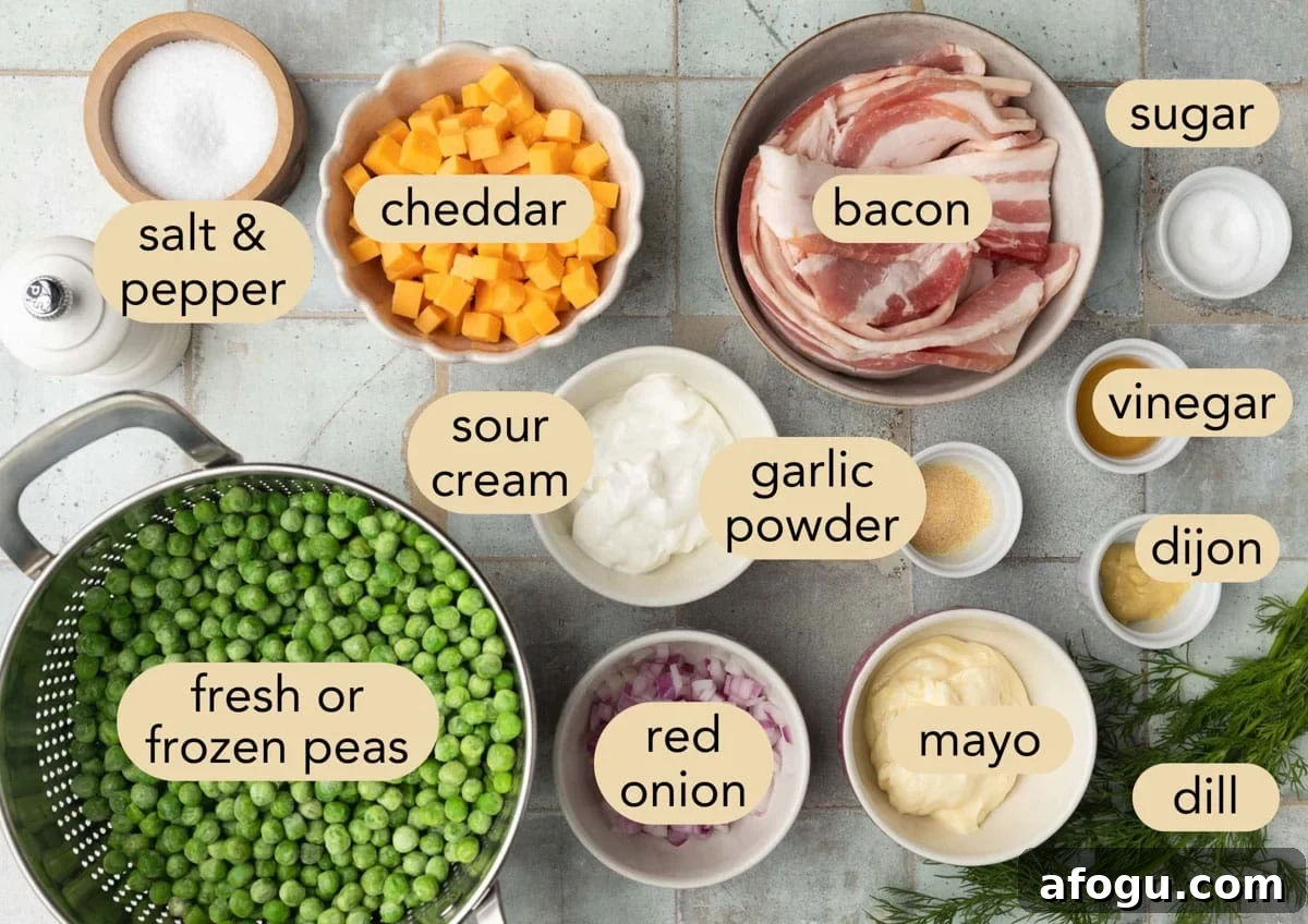 Ingredient shot for creamy pea salad arranged on a blue-gray tiled surface. The labeled ingredients include fresh or frozen peas, bacon, cheddar cheese, mayo, sour cream, red onion, dill, Dijon mustard, vinegar, garlic powder, sugar, and salt and pepper