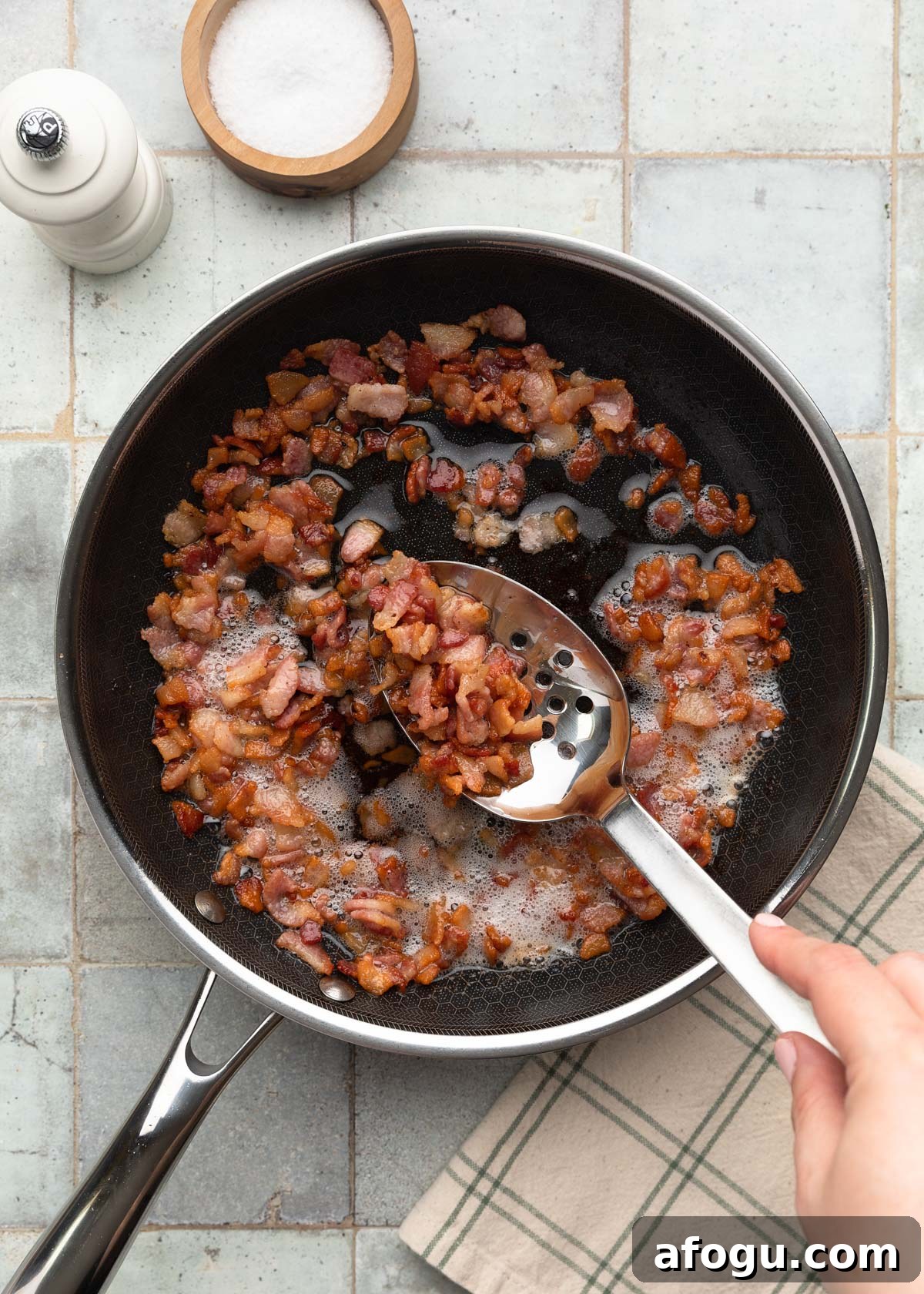 Top-down process photo of chopped bacon cooking in a skillet, with a slotted spoon lifting up crispy browned pieces from the rendered bacon fat.