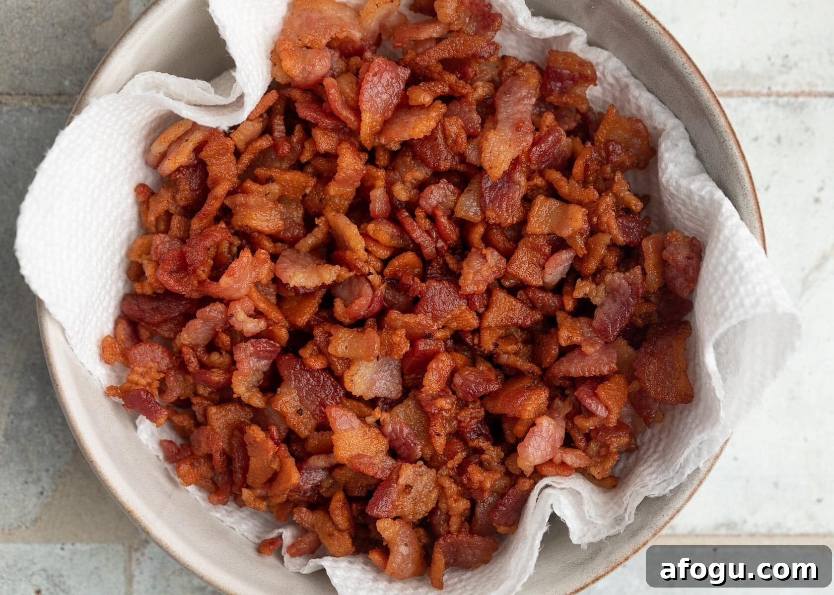 Top-down photo of crispy chopped bacon draining on a paper towel-lined bowl after cooking.
