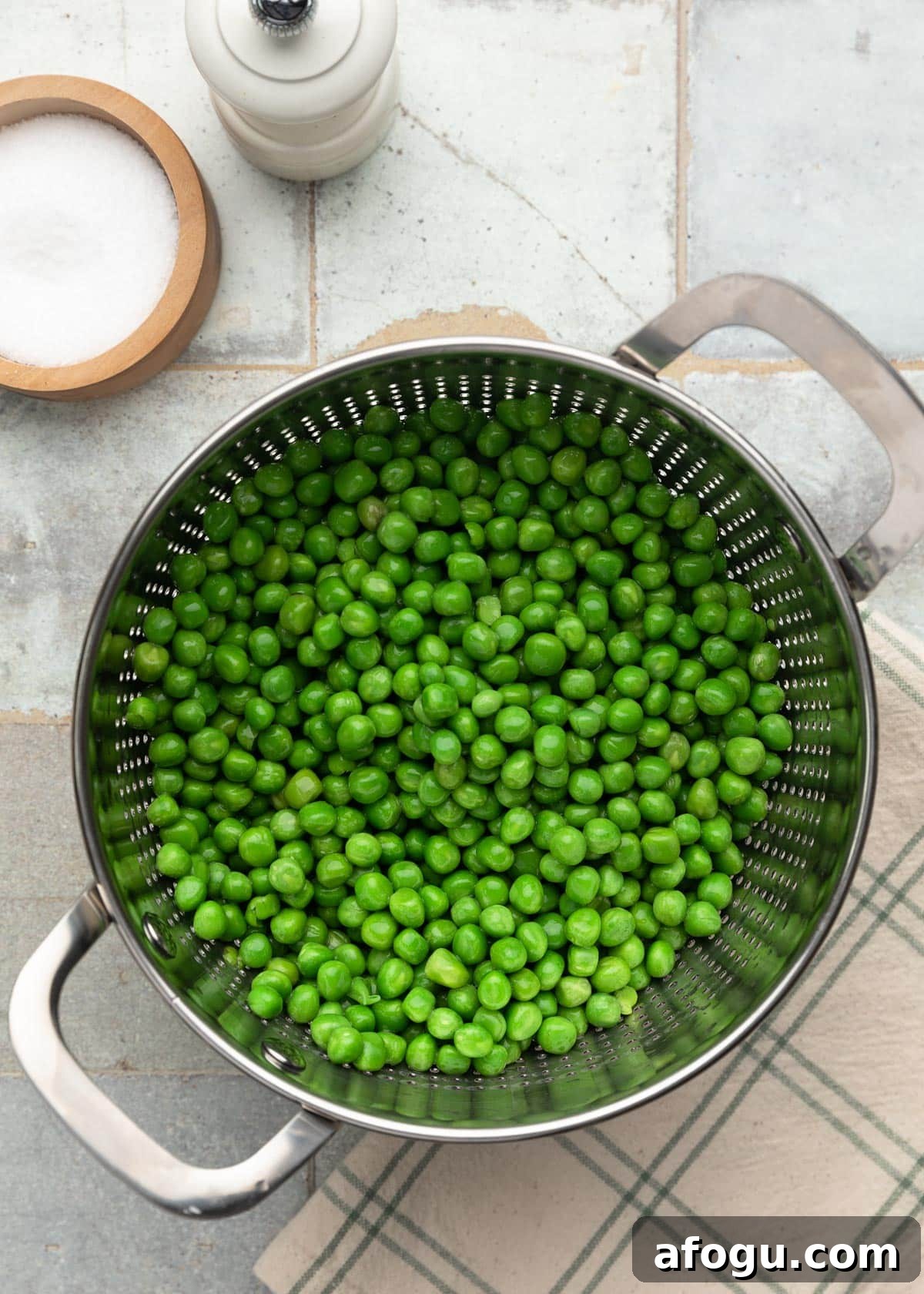 Top-down photo of bright green peas in a metal colander after blanching, set on a tiled surface with salt and pepper nearby.