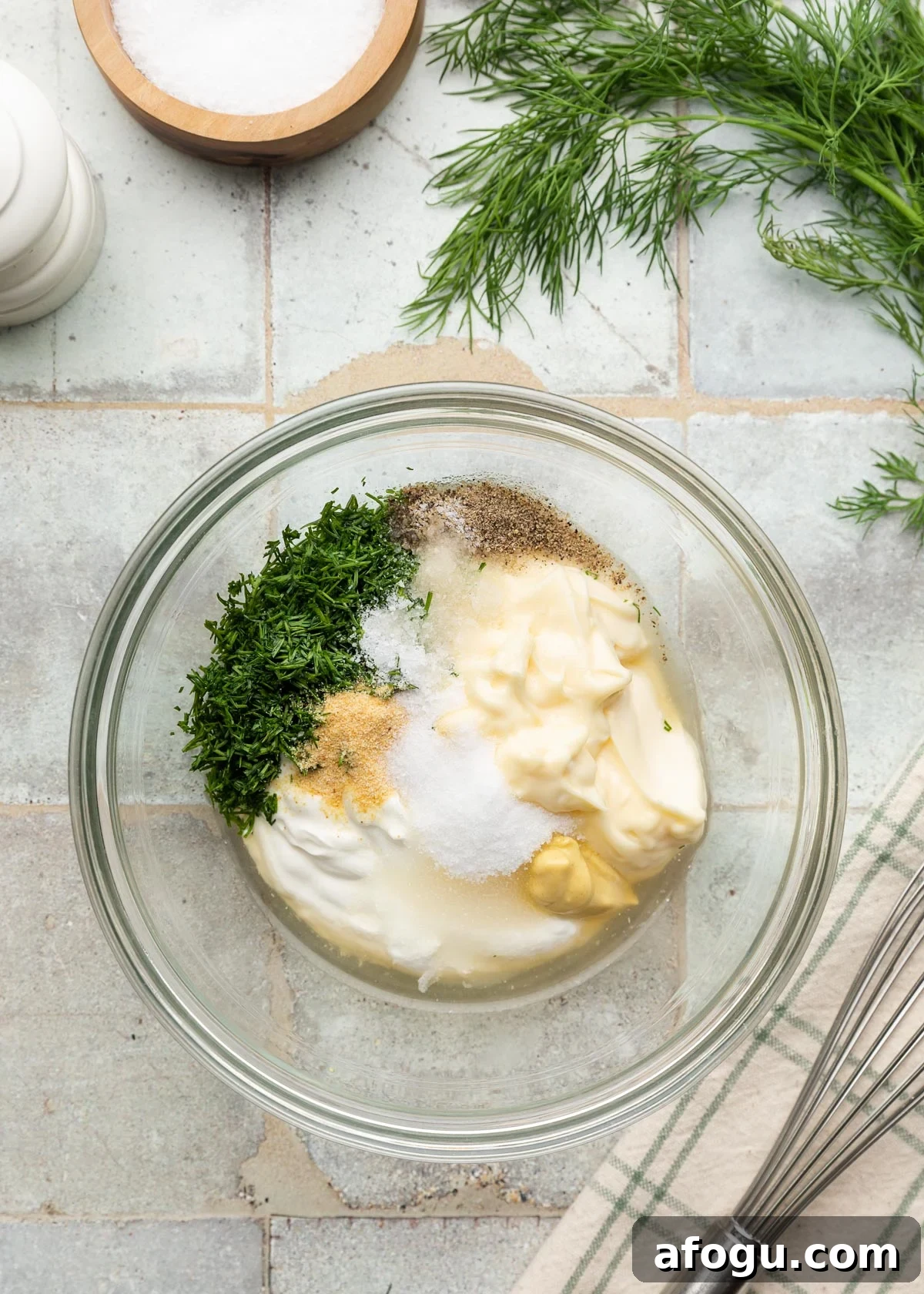 Top-down photo of ingredients for a creamy dill dressing in a glass mixing bowl, including mayonnaise, sour cream, Dijon mustard, vinegar, chopped fresh dill, sugar, garlic powder, salt, and black pepper. A whisk, salt cellar, and fresh dill are arranged around the bowl on a tiled surface.