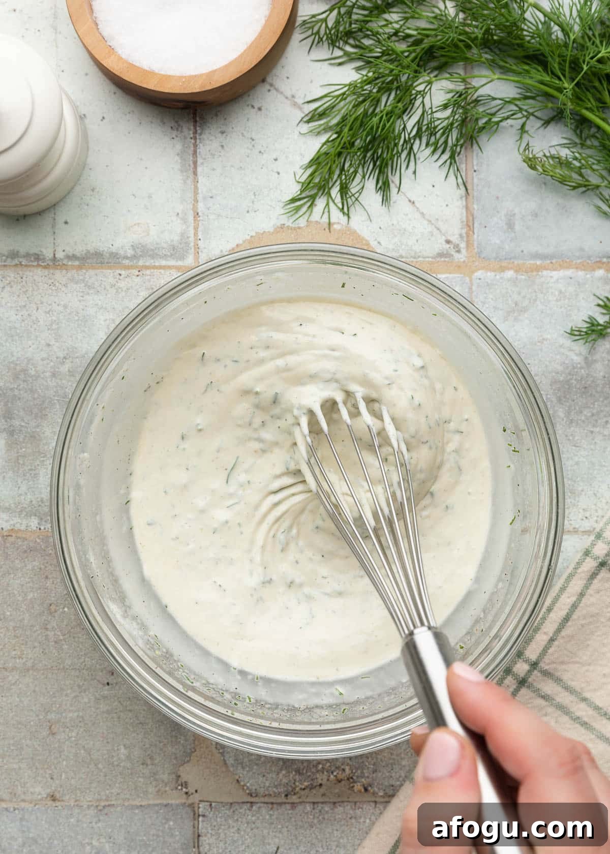 Top-down process photo of a creamy dill dressing being whisked together in a glass mixing bowl, with fresh dill and seasonings on the tiled surface nearby.