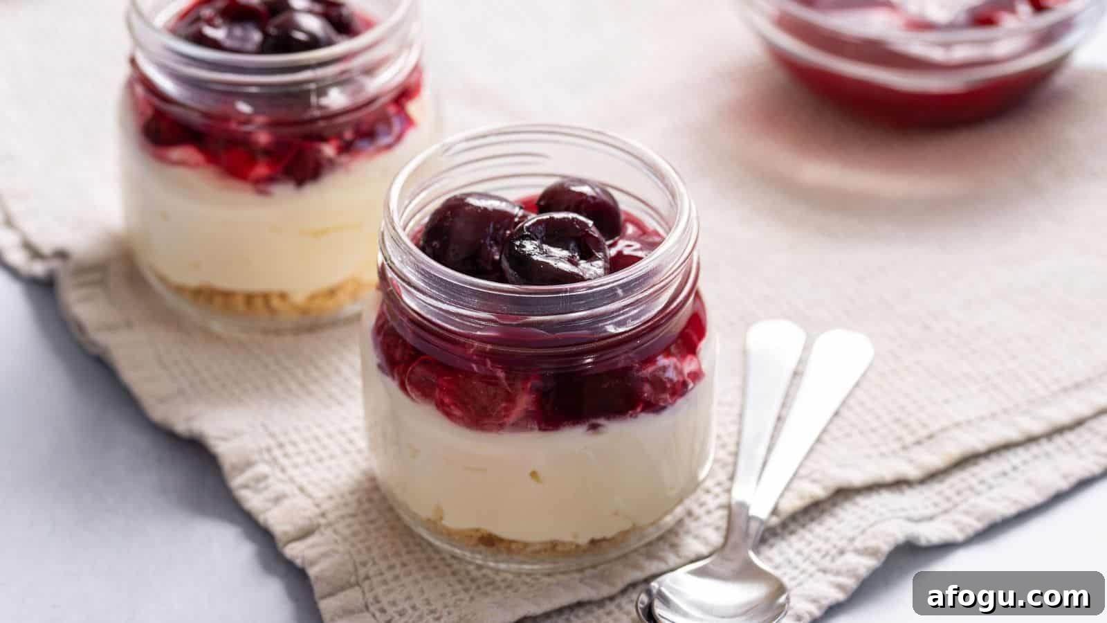 Two jars of layered cheesecake with a graham cracker base, cream cheese filling, and a topping of dark red cherries, on a cloth with spoons beside them.
