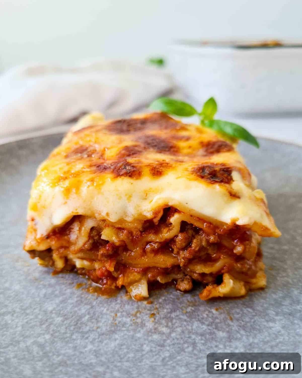 Side front on view of a slice of lasagna on a grey plate with emphasis of a thick layer of bechamel sauce.