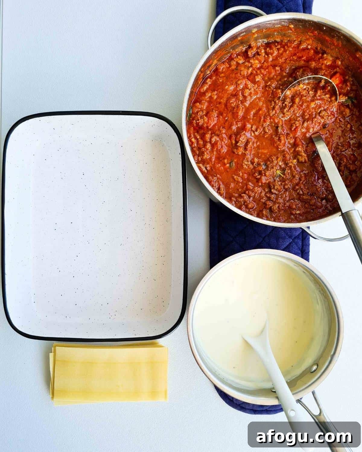 A laid out work station with a pot of cooked beef bolognese, a pot of bechamel sauce, a oven dish and a stack of dried lasagne sheets
