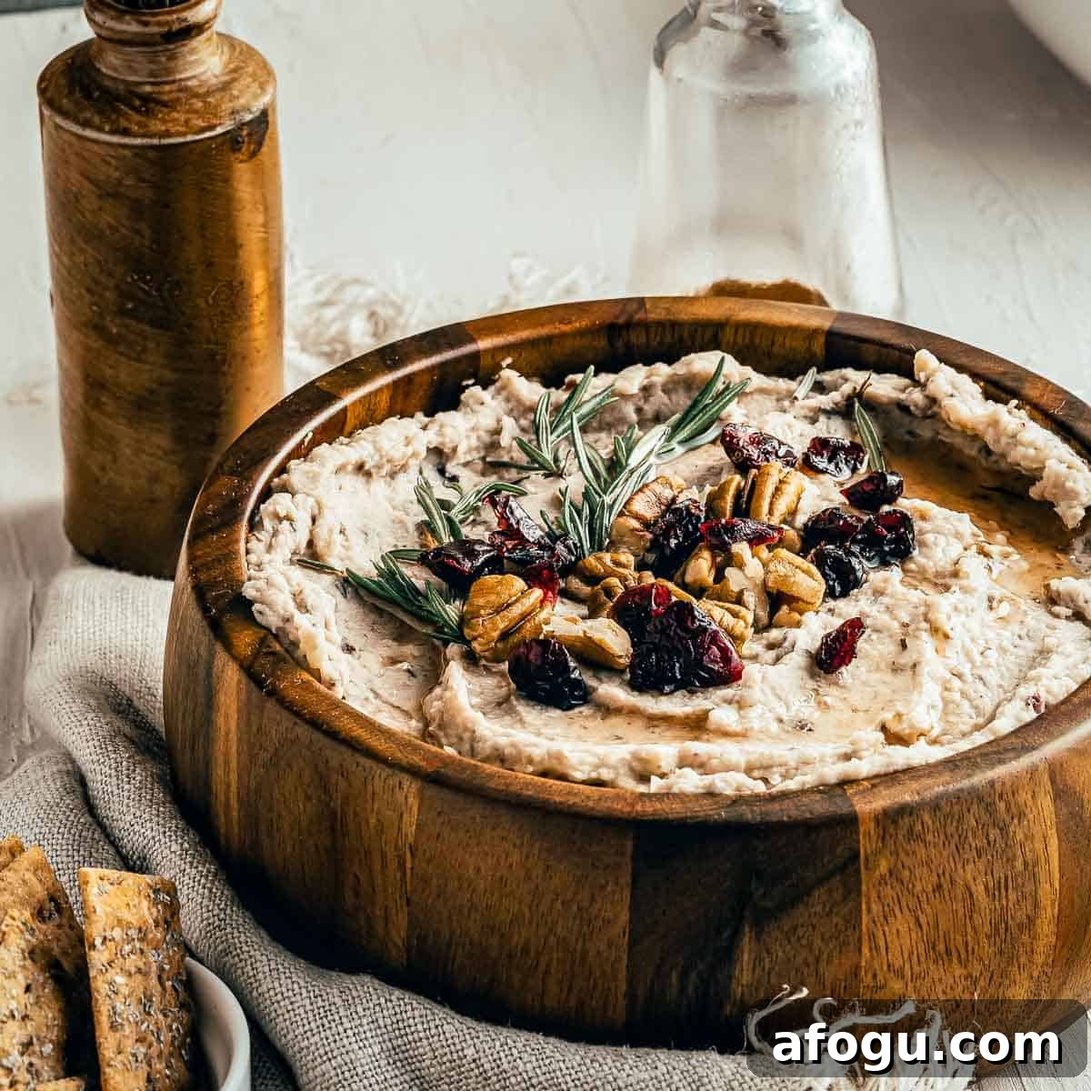 Creamy White Bean and Cranberry Hummus Recipe 5 Side shot of hummus garnished with rosemary, pecans, and cranberries in a large wooden bowl on a burlap cloth with crackers, and two jars in the background.