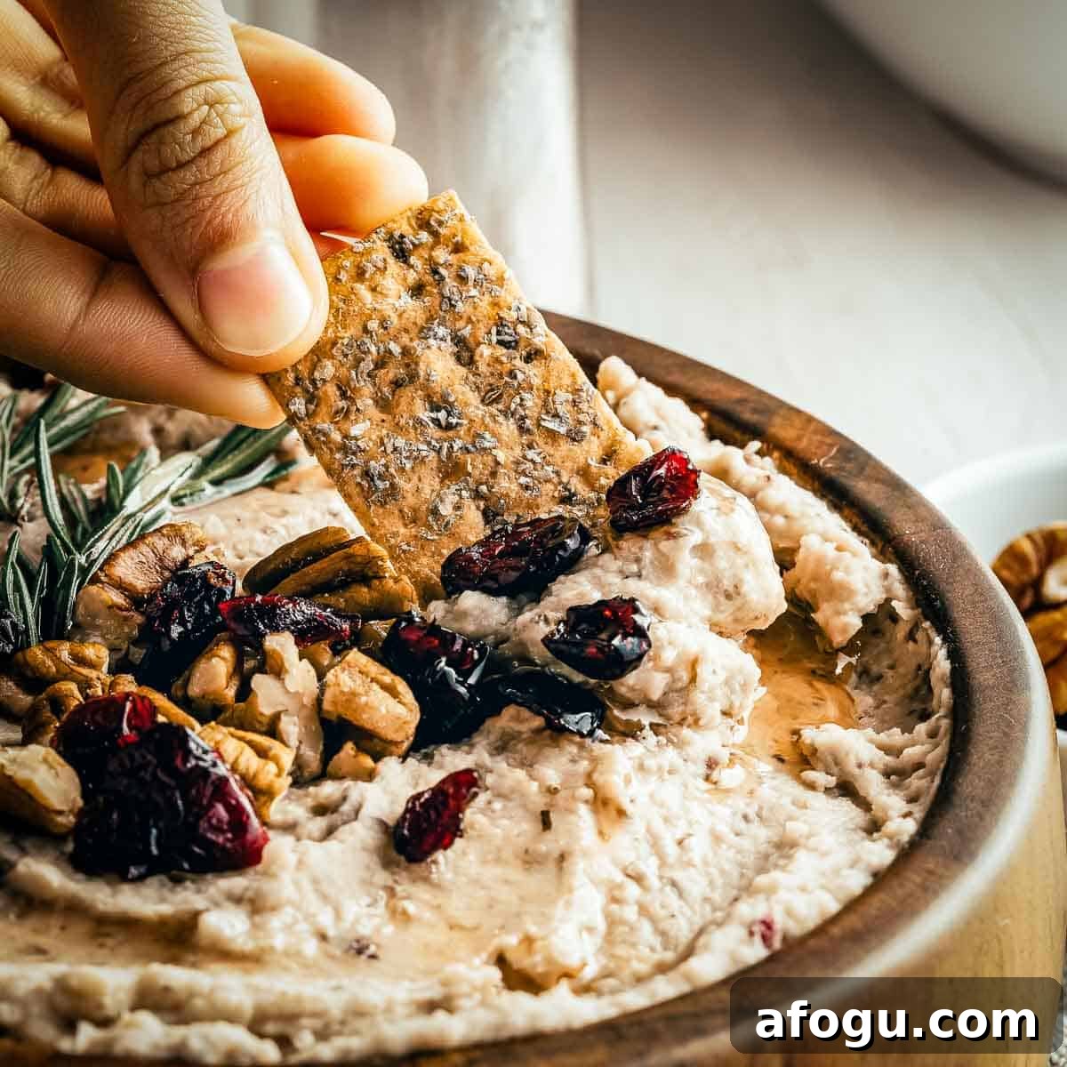 Creamy White Bean and Cranberry Hummus Recipe 6 Side close up of a hand scooping some hummus with a cracker - the hummus is garnished with rosemary, cranberries, and pecans.