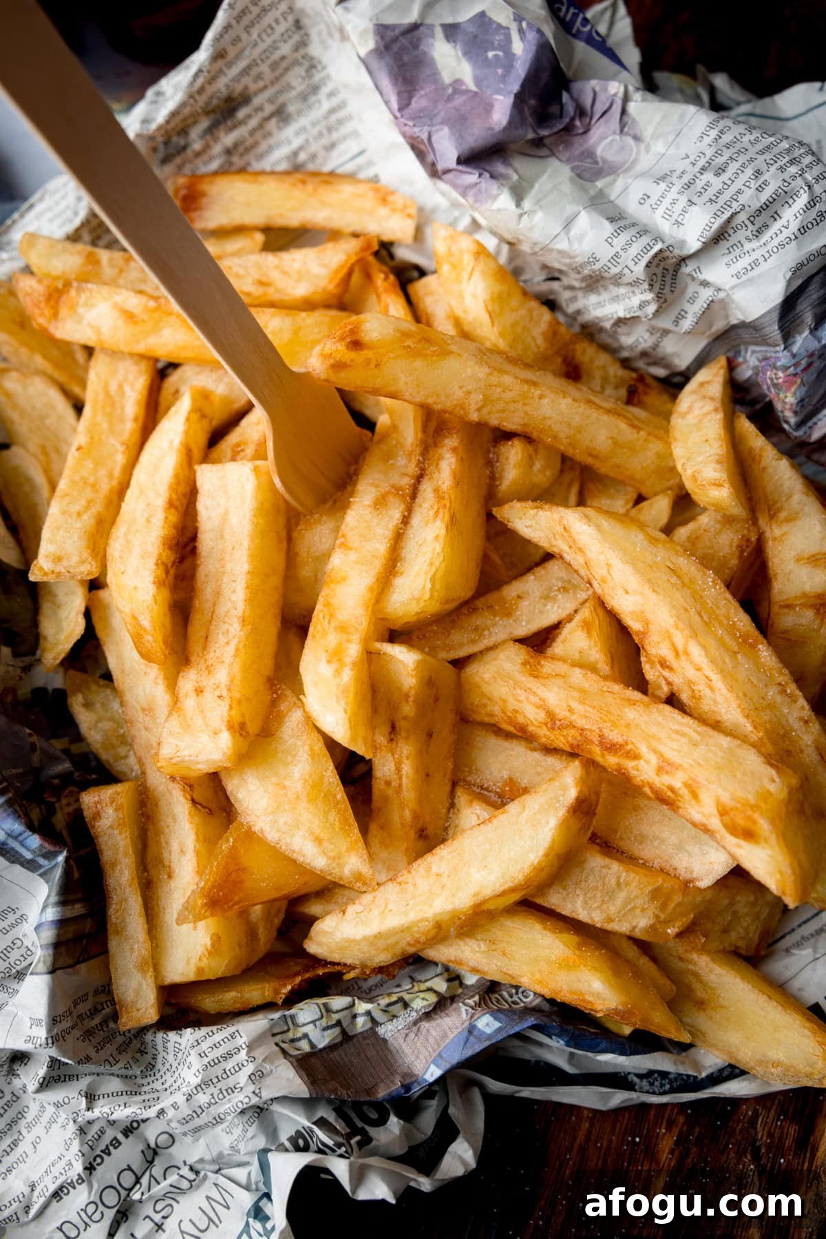 A tall, overhead image of Chip Shop-Style Chips on top of some crumpled newspaper, with a small wooden fork sticking out of the chips. The chips and newspaper are placed on a wooden surface.