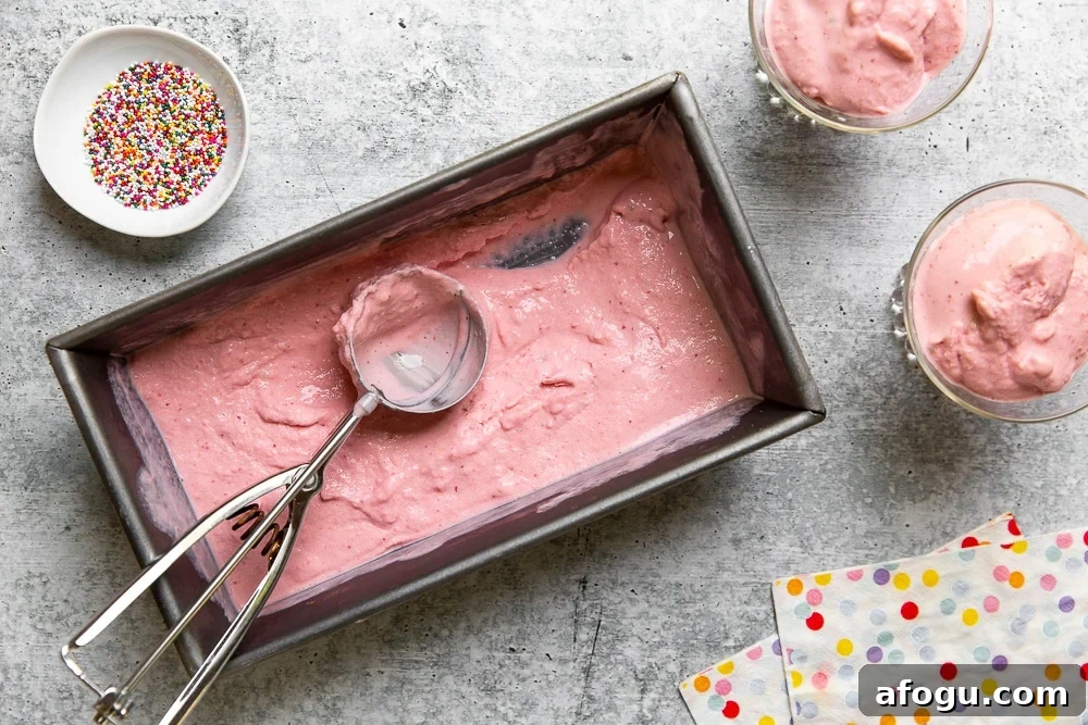 Overhead shot of the strawberry frozen yogurt in a loaf pan with an ice cream scoop, with two bowls of frozen yogurt alongside.
