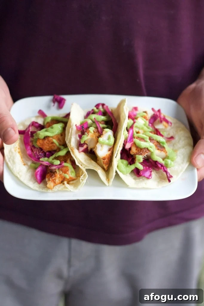 A man holding a plate with three blackened fish tacos with red cabbage and avocado crema.