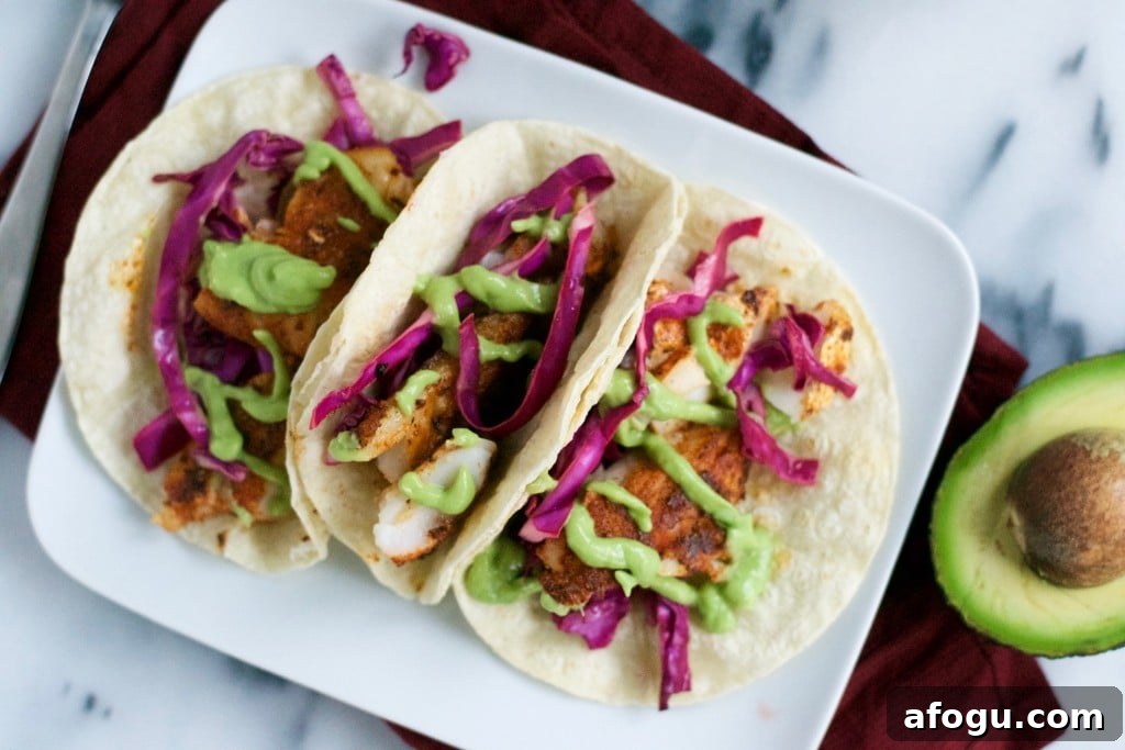 A platter of blackened fish tacos topped with red cabbage and avocado crema with a sliced avocado on the surface.