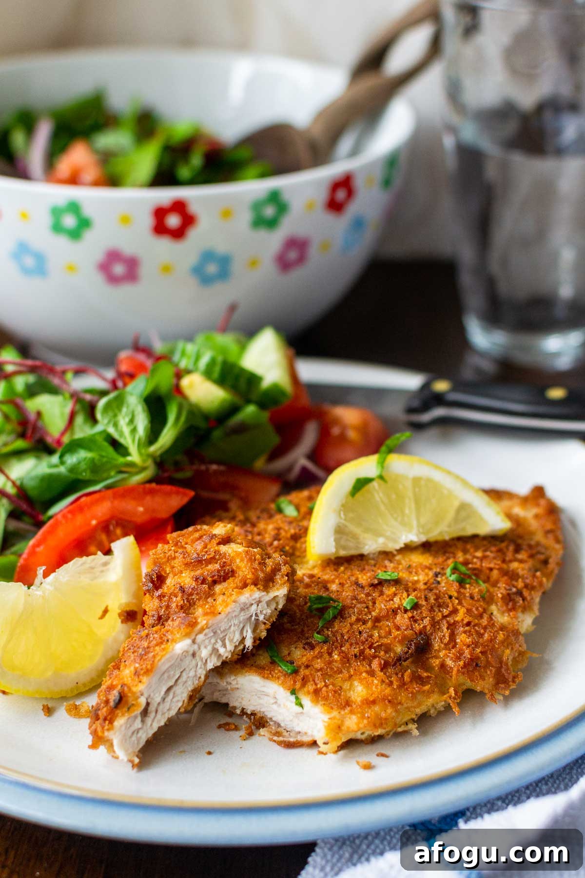 Crispy Panko Chicken with Dijon Mustard and Parmesan 2 A piece of panko chicken with the end sliced off to reveal inside on a white plate with blue rim, with brightly colored salad bowl and water glass in background.