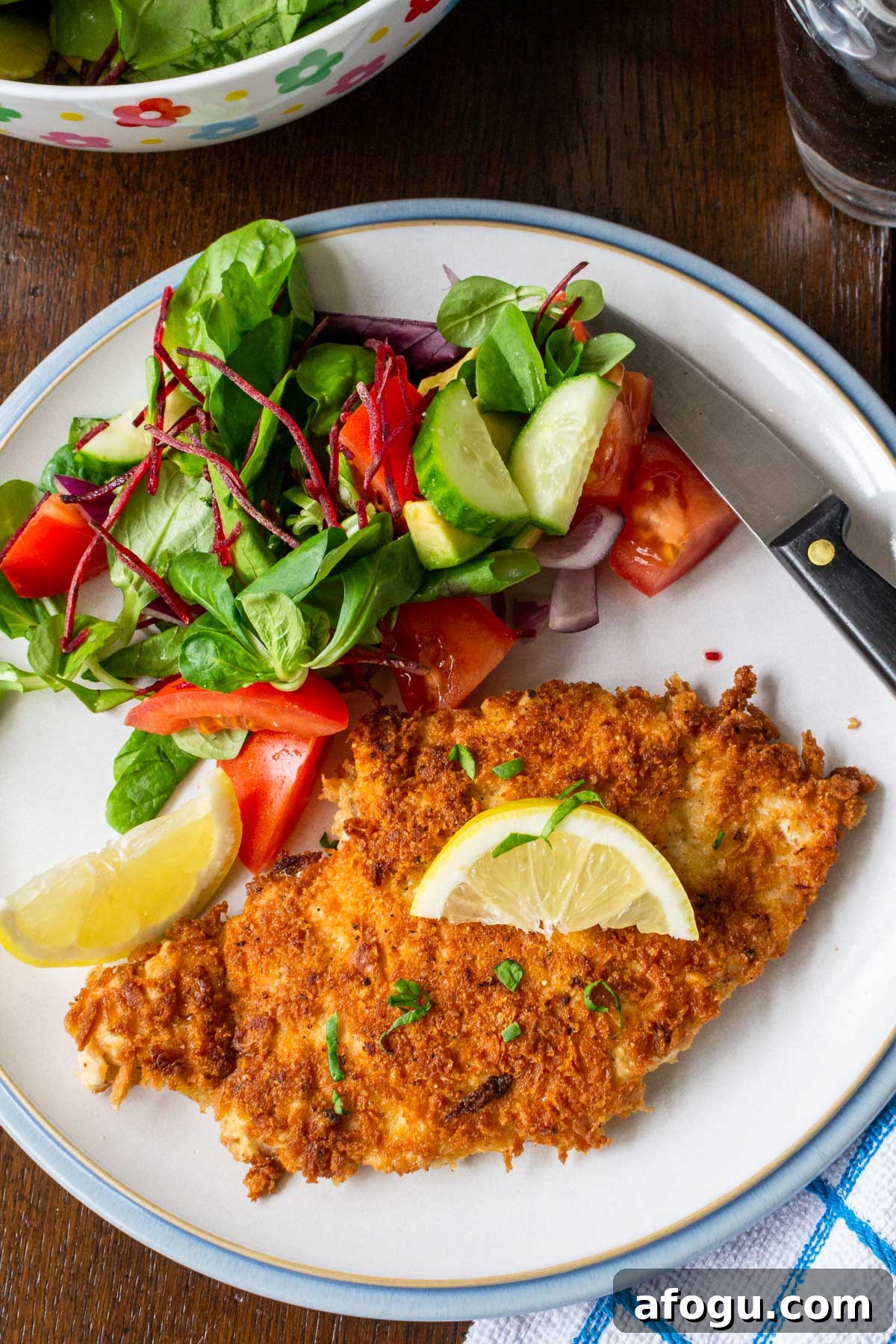Crispy Panko Chicken with Dijon Mustard and Parmesan 7 A crispy looking panko breadcrumbs crusted chicken breast on a plate with a blue rim from above with a simple side salad on the plate and with blue and white checked tea towel below on a wooden table.