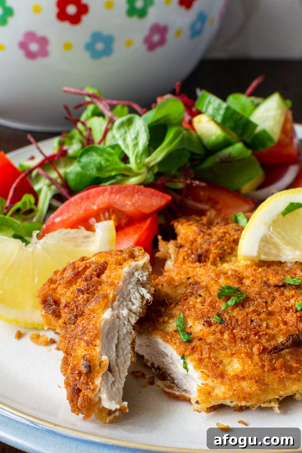 Crispy Panko Chicken with Dijon Mustard and Parmesan 8 Closeup view of the inside of a piece of panko chicken breast on a white plate with blue rim with a simple salad on the plate in the background, plus salad bowl.