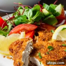 Crispy Panko Chicken with Dijon Mustard and Parmesan 9 Closeup view of the inside of a piece of panko chicken breast on a white plate with blue rim with a simple salad on the plate in the background, plus salad bowl.