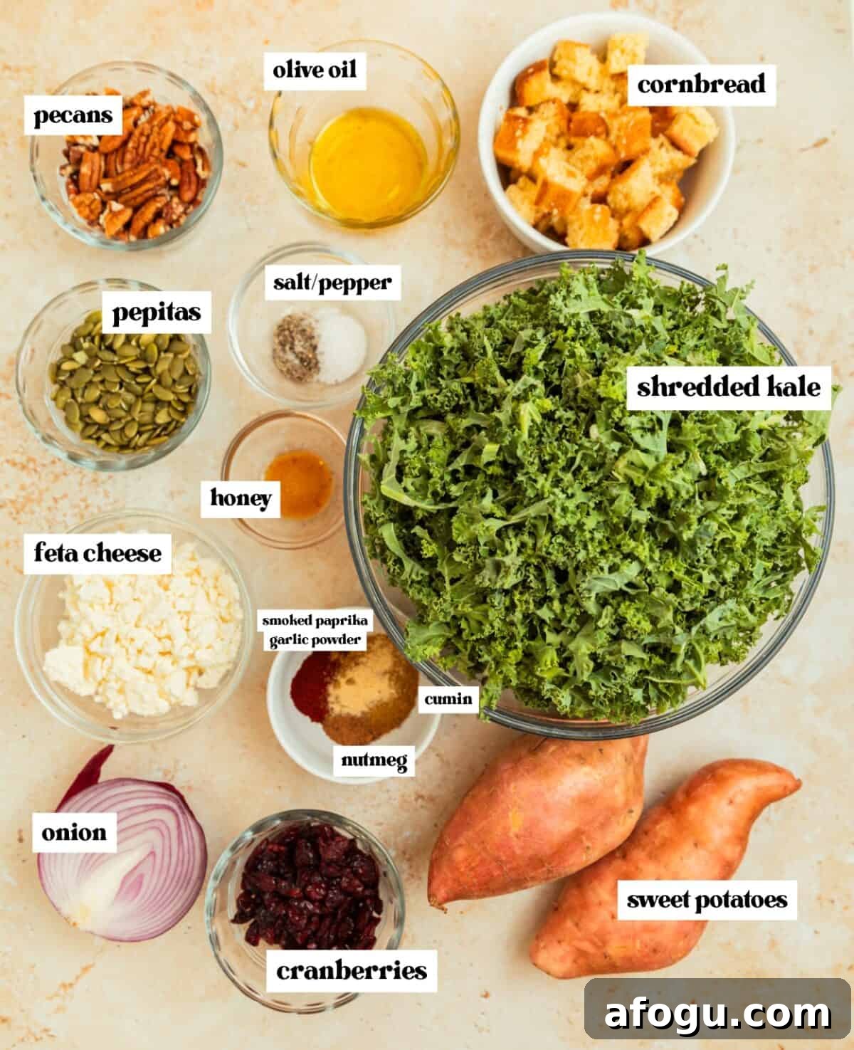 Shredded kale in glass bowl, cornbread cubes, feta, sweet potatoes, and other ingredients on surface. 