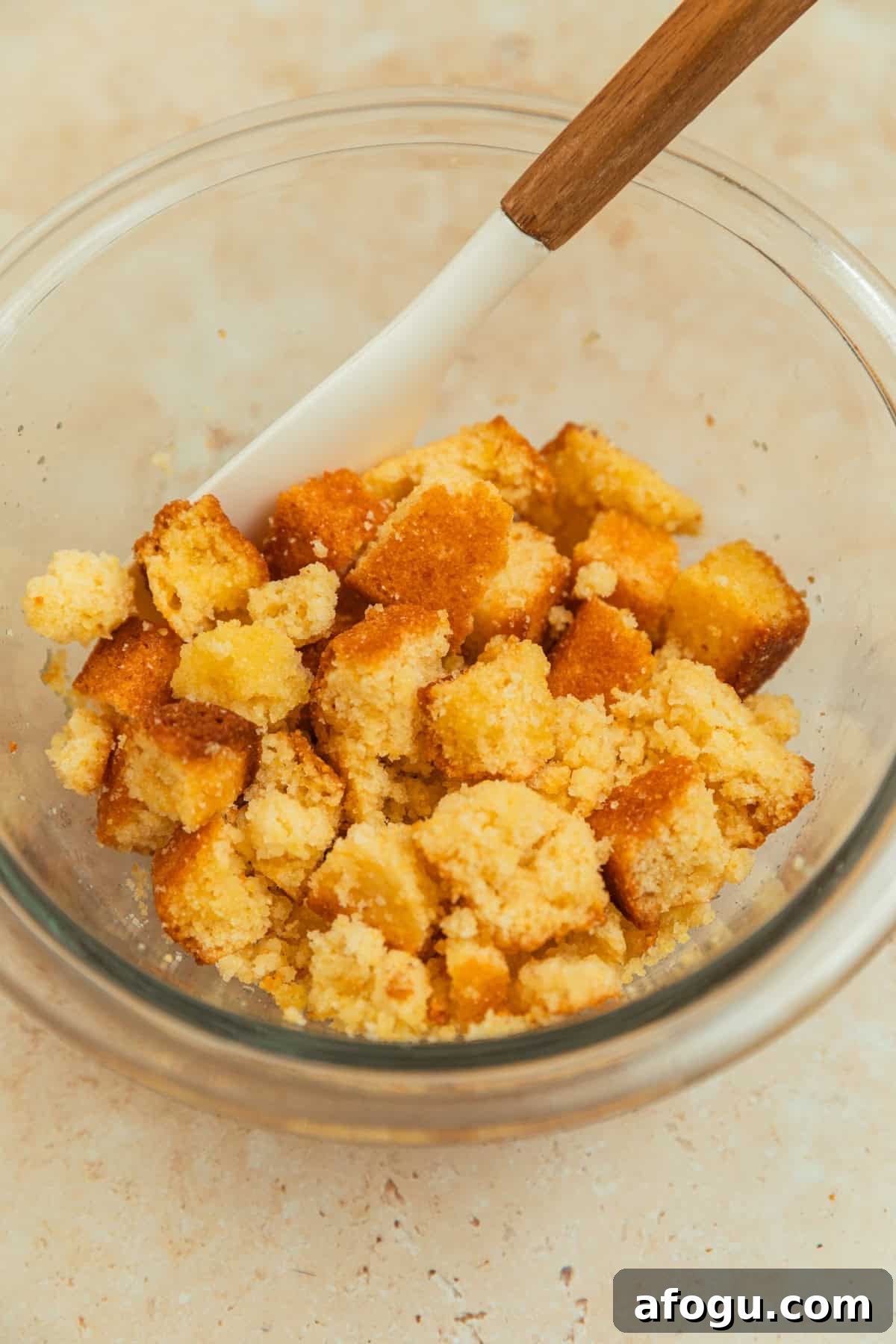 Cornbread cubed in glass mixing bowl with oil and spatula.