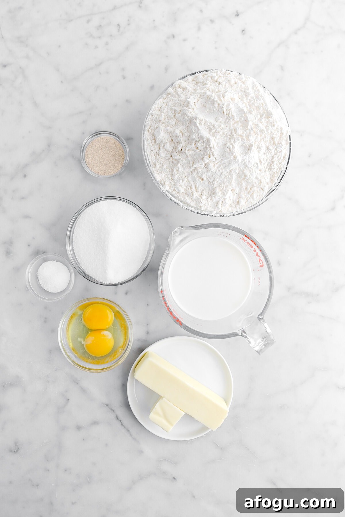 yeast, flour, sugar, salt, milk, eggs, and butter on marble surface.