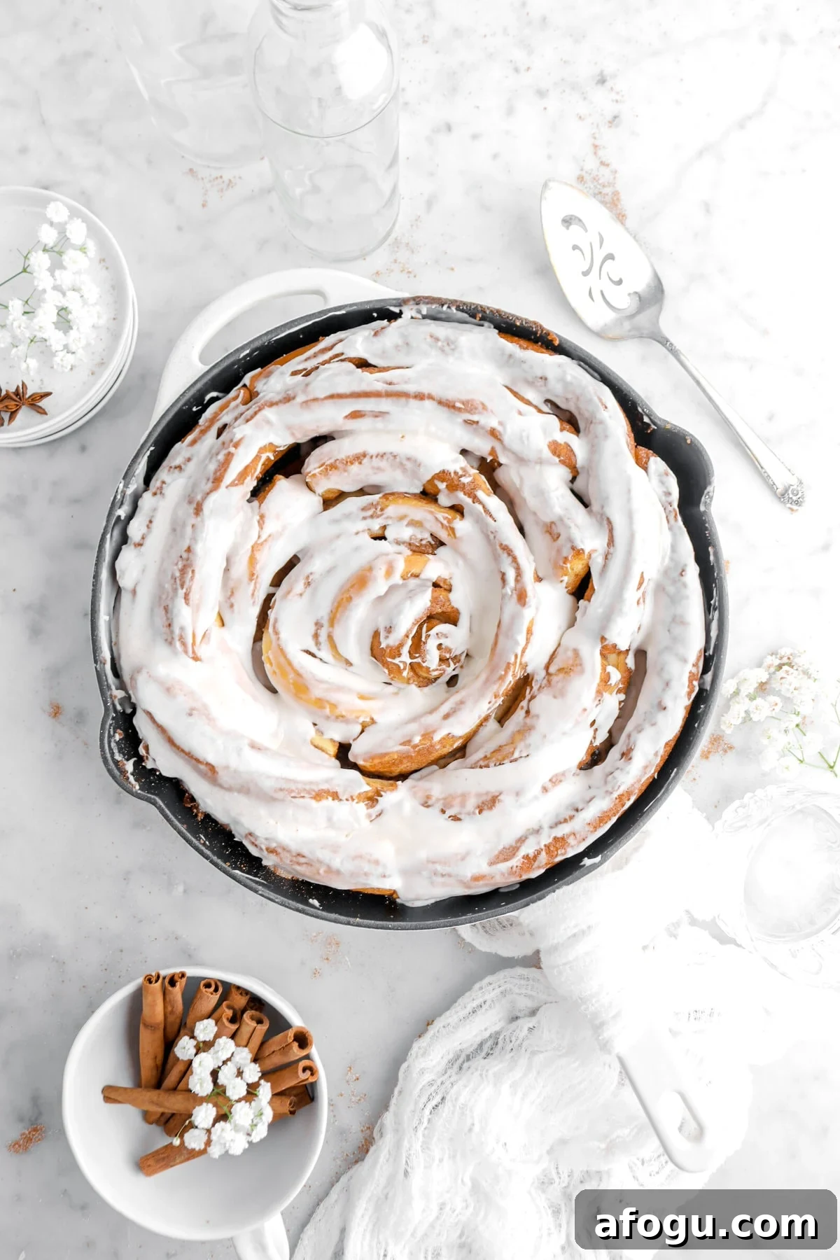 frosted giant cinnamon roll in skillet on marble surface with cinnamon sticks and flowers around.