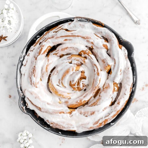 overhead image of iced cinnamon roll in skillet with white flowers, cinnamon sticks, and a white cheesecloth around.