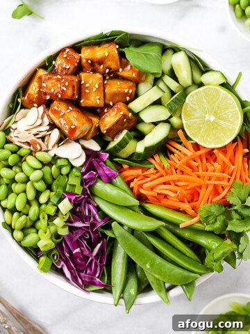 A white bowl holding a large salad, consisting of: sesame tofu, cucumbers, a lime, carrots, snap peas, red cabbage, edamame, and almonds.