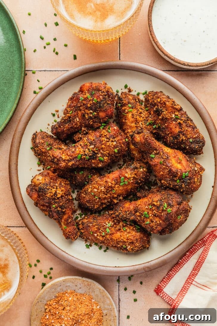 Crispy Dry-Rub Chicken Wings — Oven or Air Fryer Recipes 9 A stoneware plate of dry rub chicken wings next to glasses of beer, an orange and white striped linen, brown bowls of ranch and rub, and a green plate on an orange tile table.