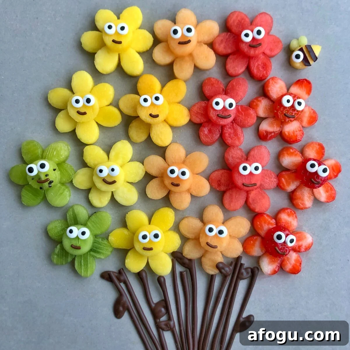 Elegant Fruit Platter Ideas for Special Occasions 2 An arrangement of colorful fruit slices shaped like flowers with cartoon faces and chocolate stems. A mango bee is seen in the background.