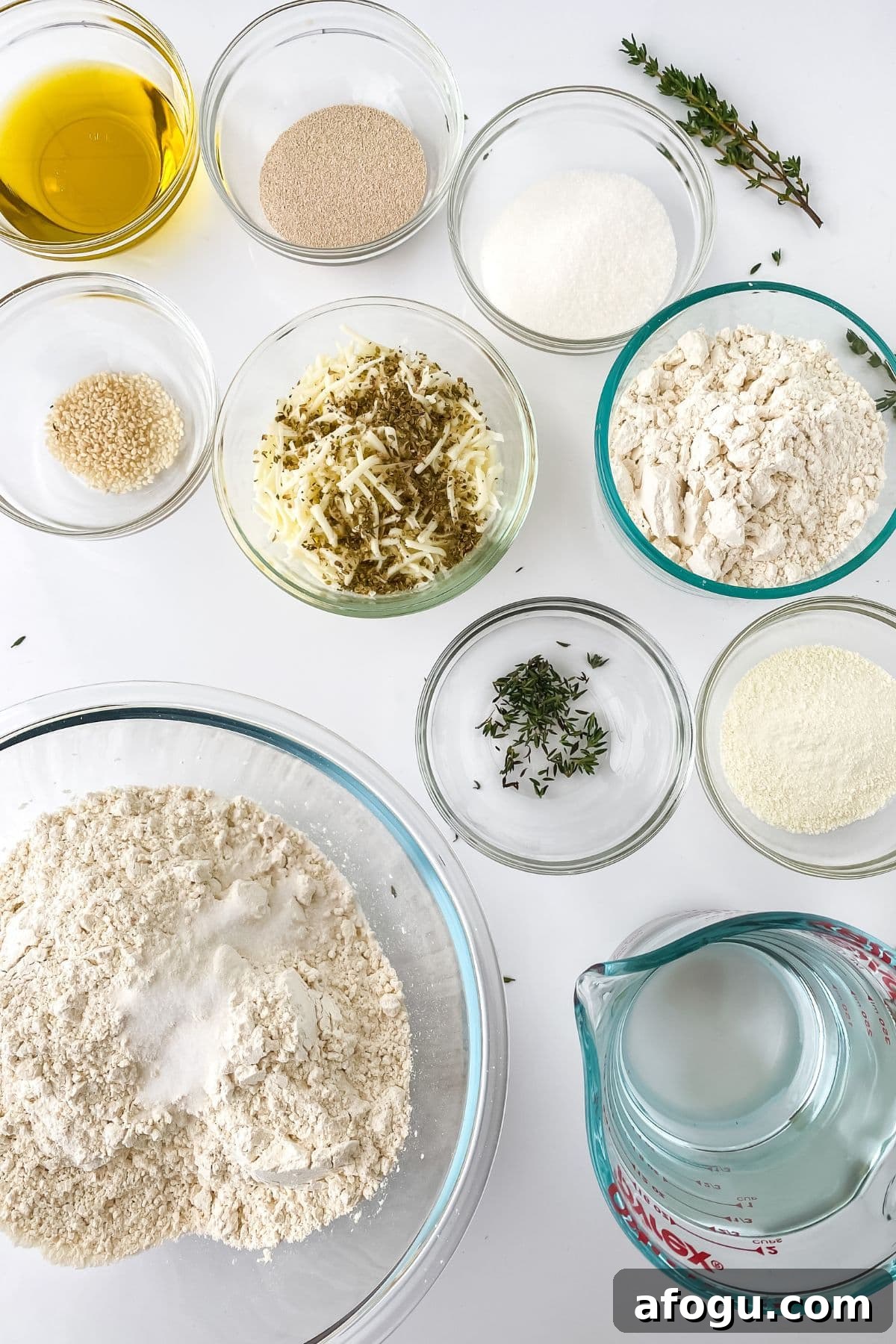 Assorted baking ingredients in glass bowls on a white surface, including flour, grated cheese, yeast, sugar, oil, herbs, and water—perfect for making homemade Pull Apart Pizza Bread.