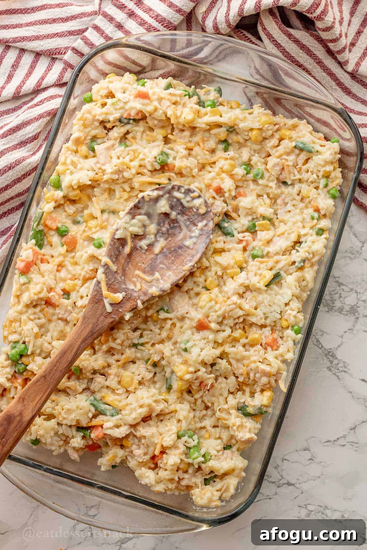 Spreading unbaked chicken and veggies and rice in glass pan with wooden spoon.