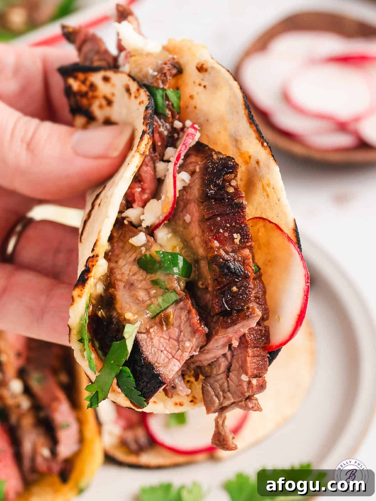 These vibrant Baja Steak Tacos are loaded with zippy marinaded grilled skirt steak and topped with cotija cheese, radishes, and cilantro. A hand holding a Baja street taco up to the camera.