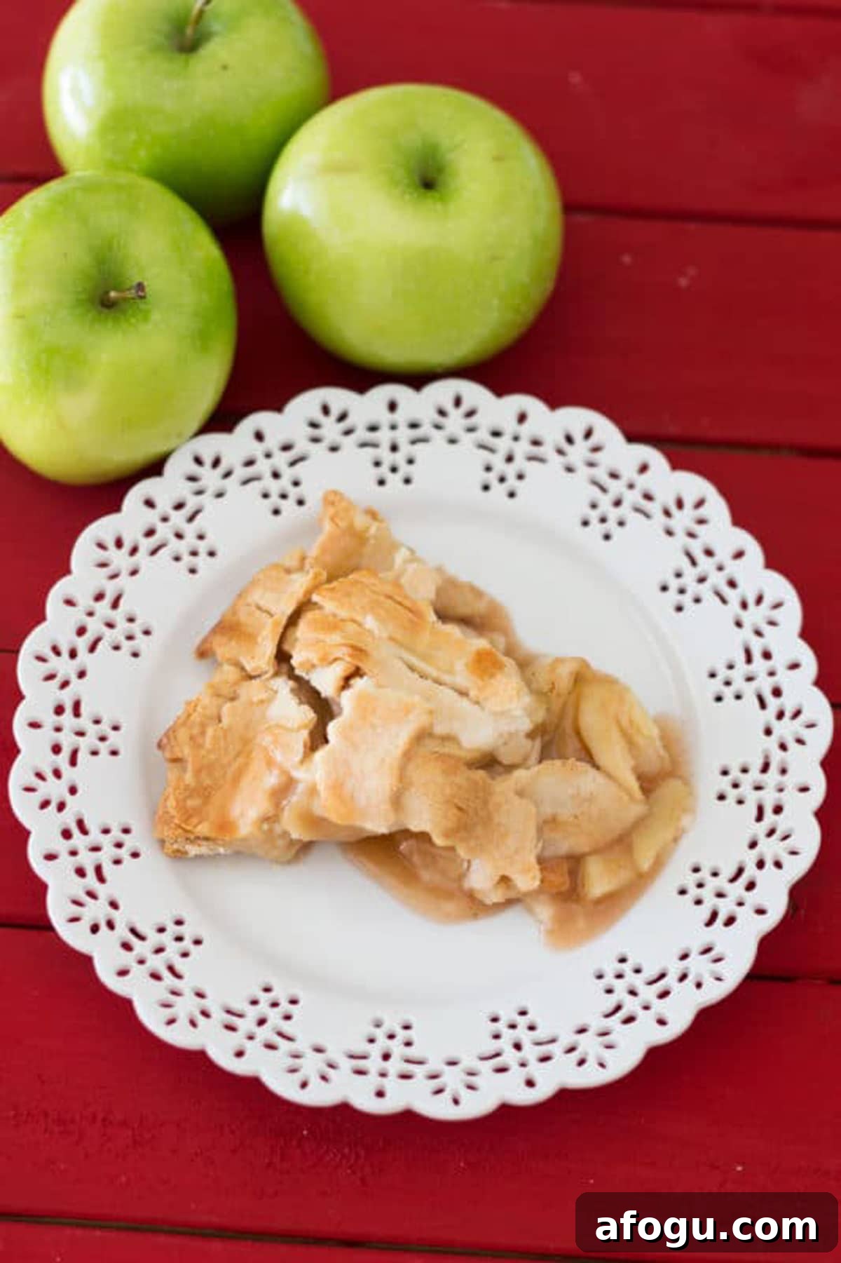 White lace place containing a piece of homemade apple pie sitting on a red table, 3 granny smith apples on table.