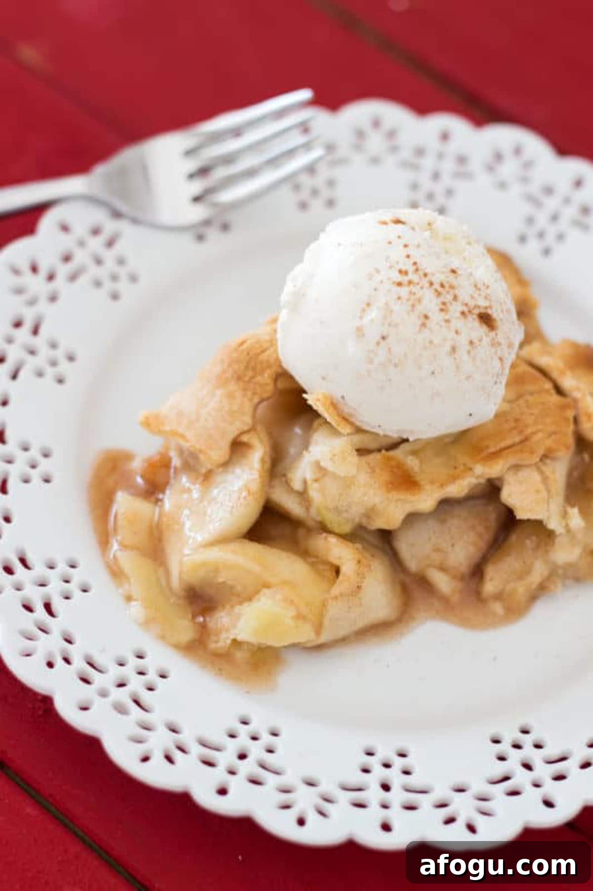 Piece of pie served on a white lace place sitting on a red table, topped with Vanilla ice cream and cinnamon.