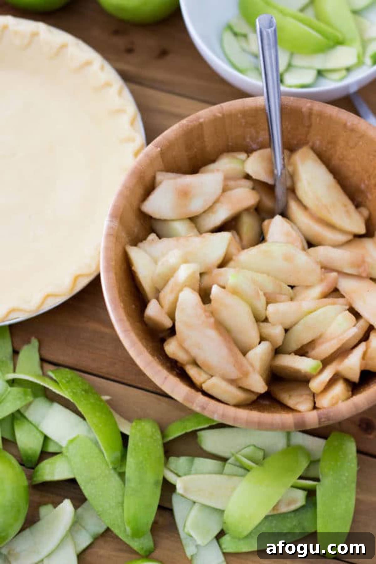 Wooden bowl containing apples covered with pie filling, pie shell and apple peelings on wooden table.