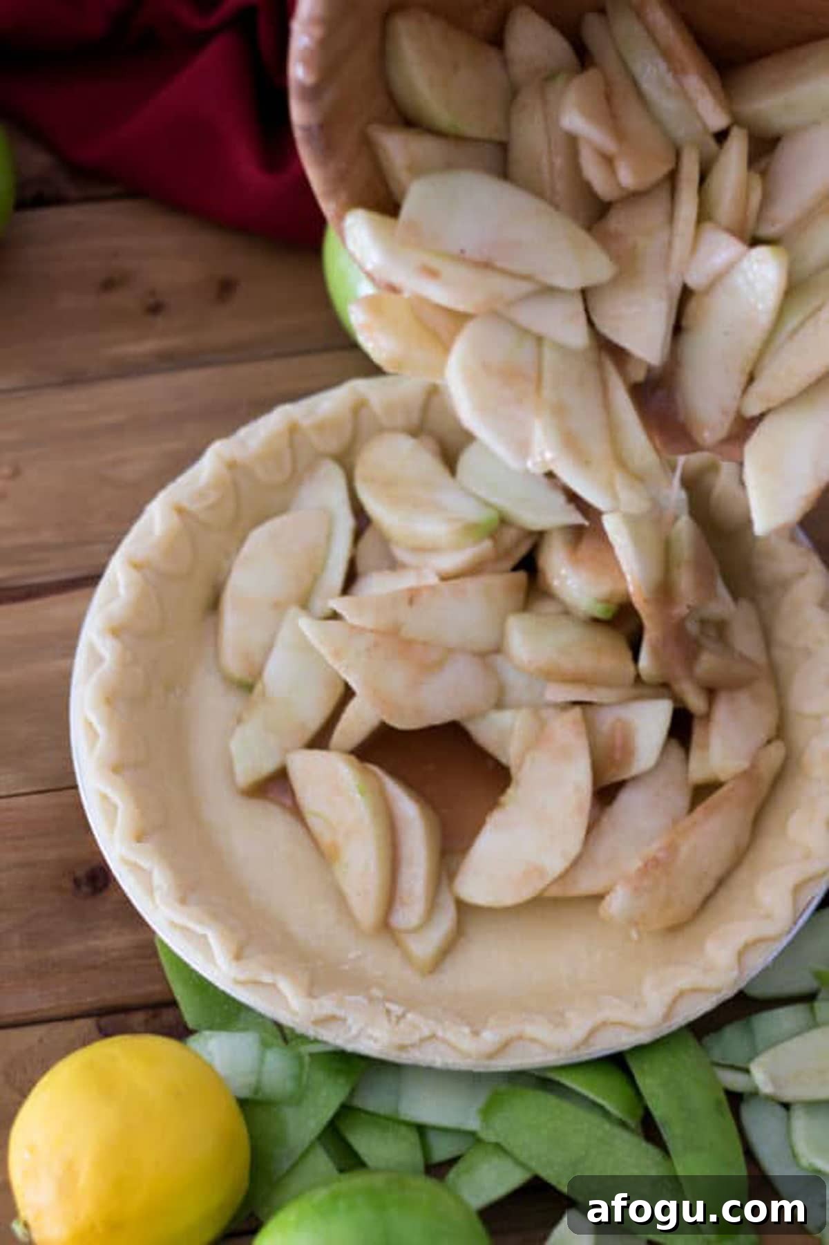 Coated apples being poured from bowl into a pie shell, lemon and apple peelings on table.
