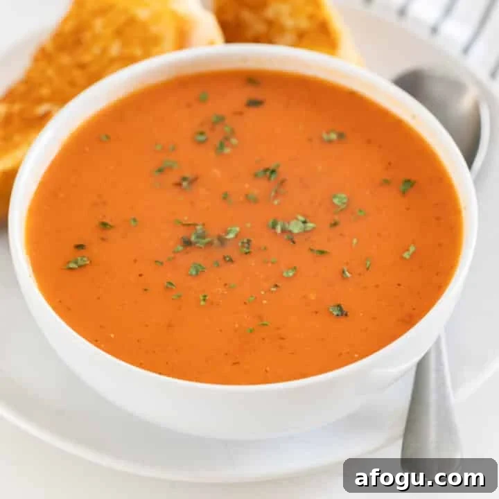 A bowl of tomato soup on a plate with a spoon and grilled cheese.