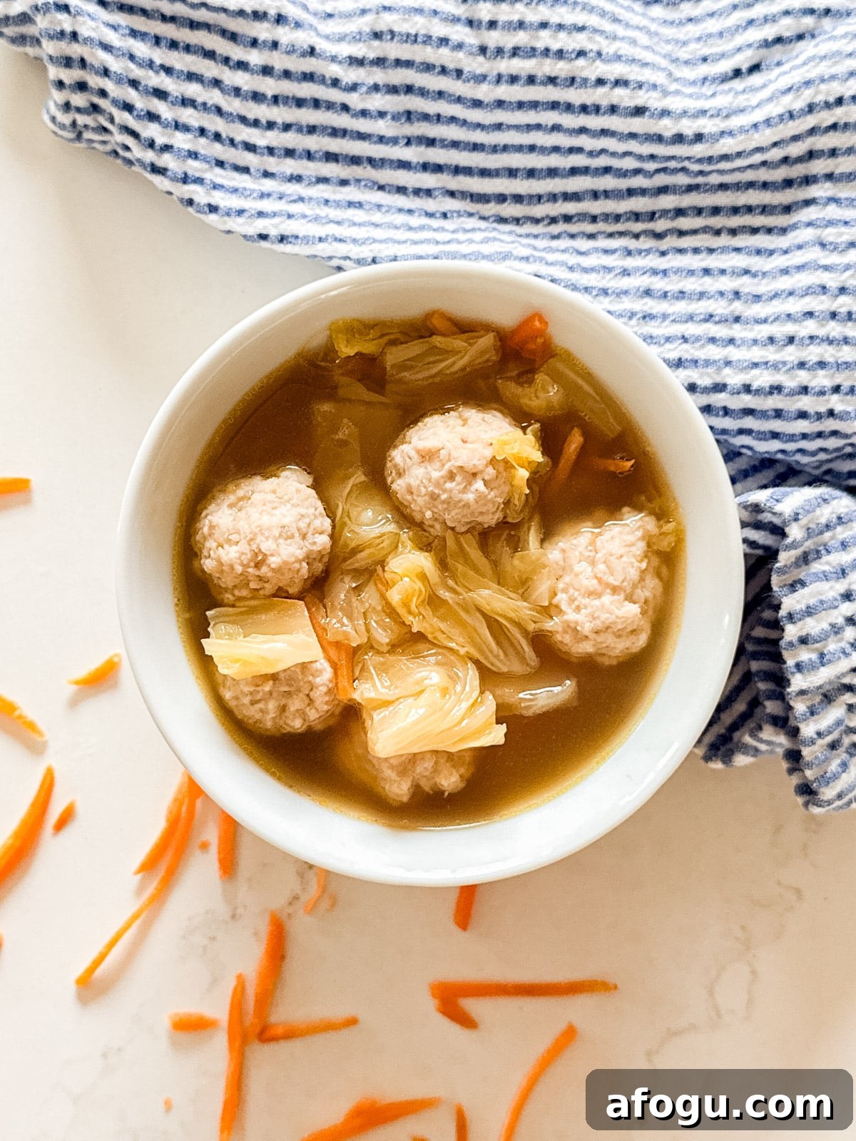 Asian Cabbage and Pork Meatball Soup Recipe 9 An overhead image of a bowl of Asian meatball soup with carrots and a striped napkin next to it.