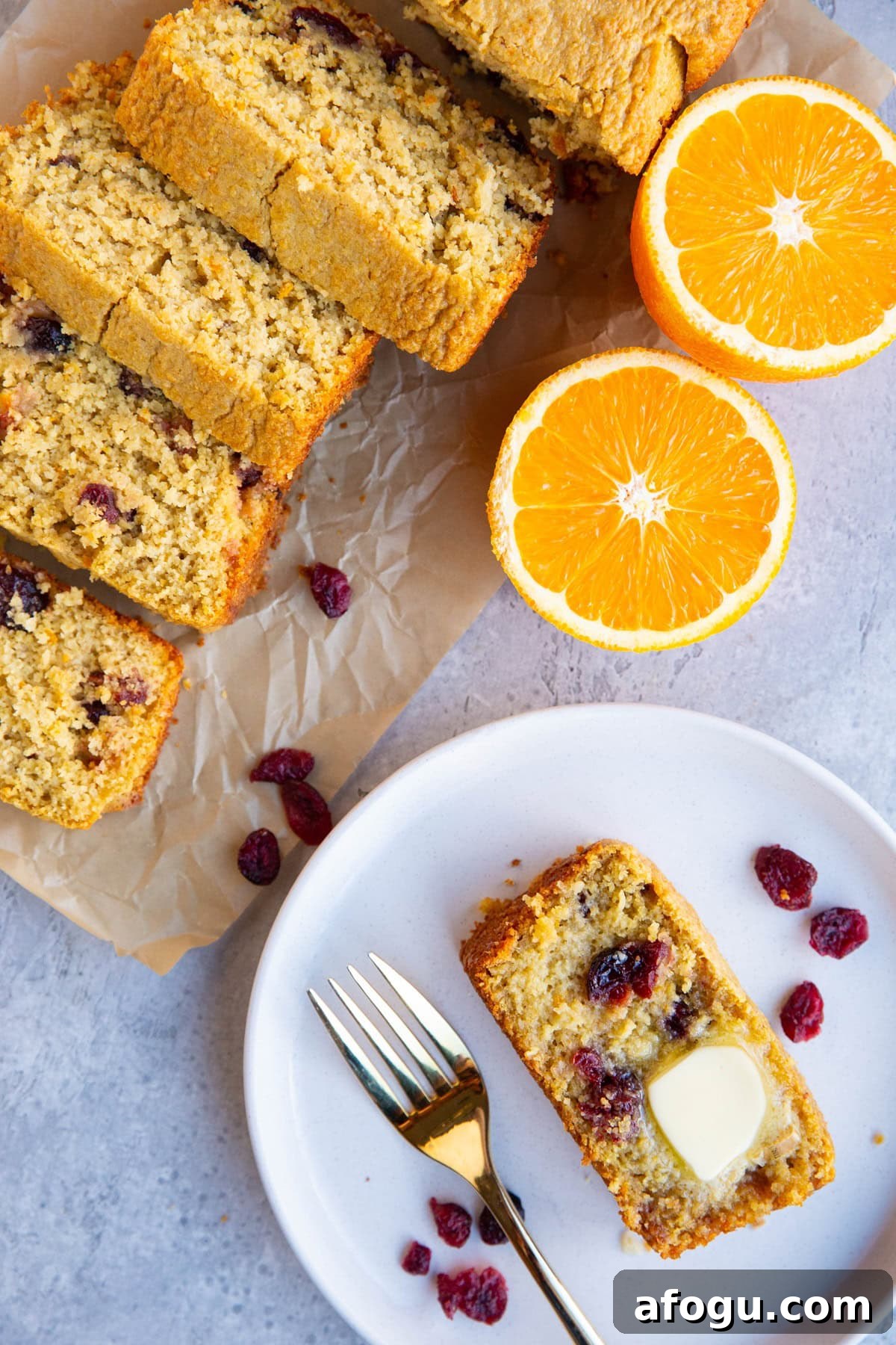 Slice of cranberry orange bread on a plate with the rest of the loaf next to it cut into slices and a fresh orange to the side.