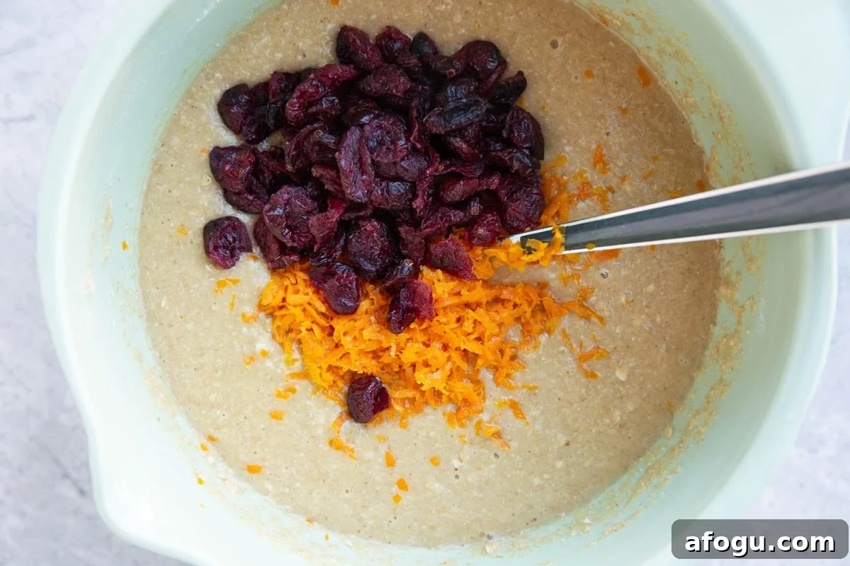 Muffin batter in a mixing bowl with orange zest and dried cranberries on top, ready to be mixed in.