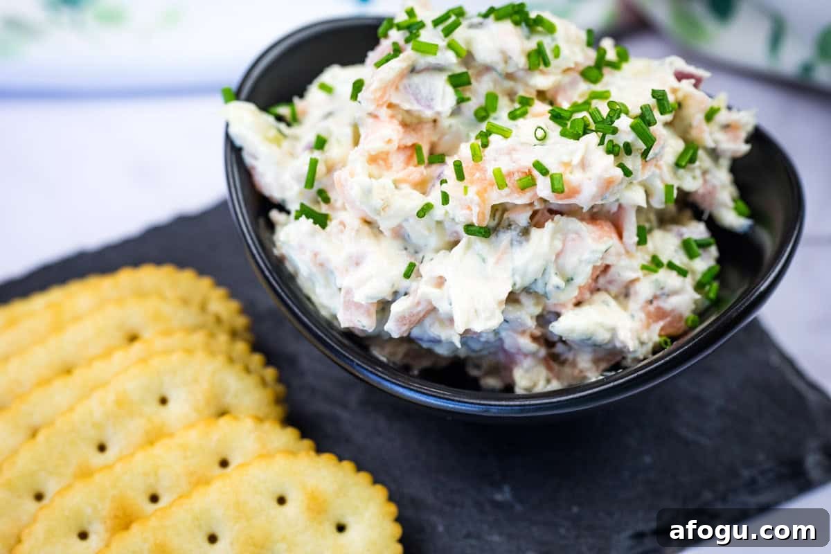 Bowl of smoked salmon spread garnished with chopped chives next to crackers on a slate platter.