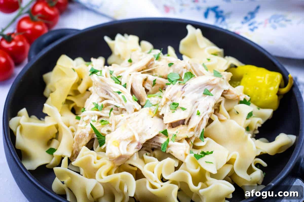 A black bowl containing egg noodles topped with Slow Cooker Mississippi Chicken and garnished with parsley, accompanied by a pickled pepper.