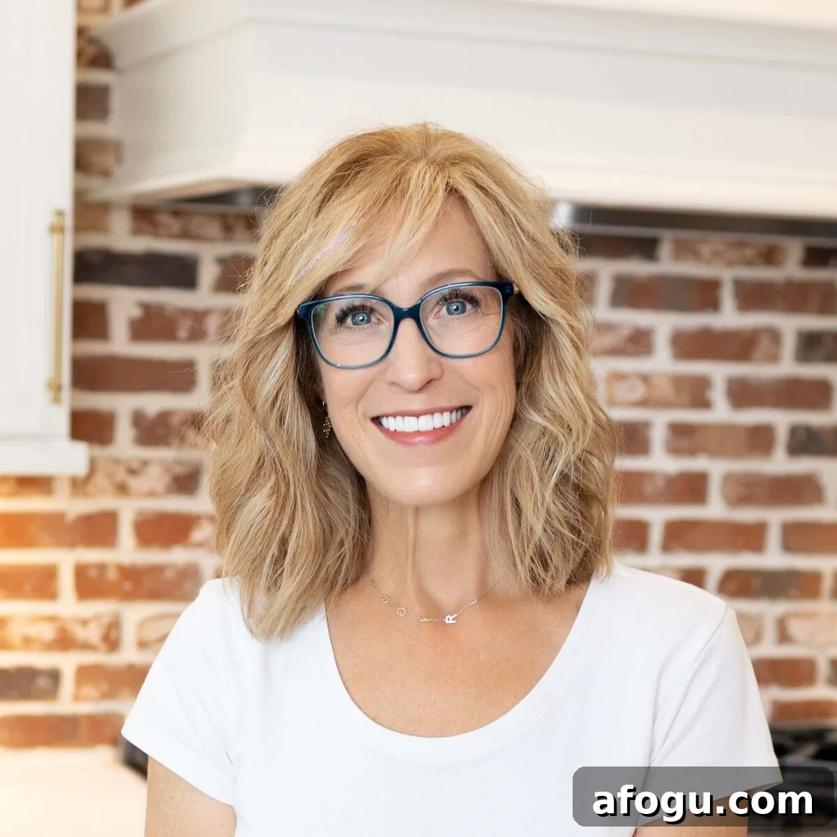 Dairy-Free Green Breakfast Smoothie Recipe 3 Person with shoulder-length blond hair and blue glasses smiling indoors in front of a red brick wall.