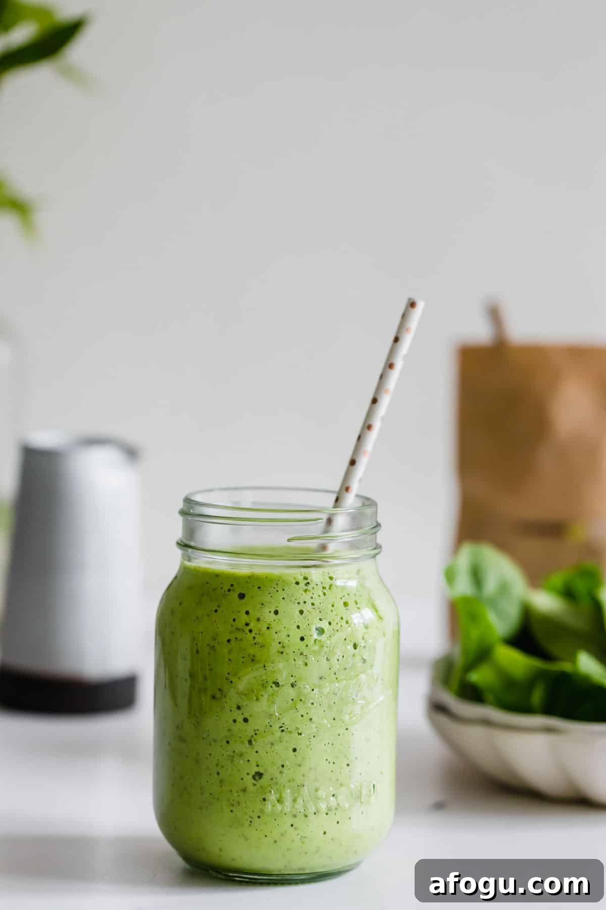 Dairy-Free Green Breakfast Smoothie Recipe 5 A green breakfast smoothie in a mason jar with a spotted straw sits on a white surface, with spinach leaves and a brown paper bag in the background.