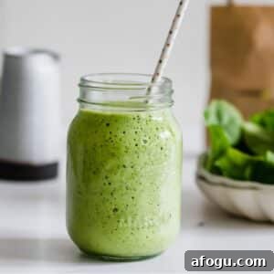 Dairy-Free Green Breakfast Smoothie Recipe 6 A mason jar filled with a green smoothie and a polka dot straw, set on a white surface with spinach leaves in the background.