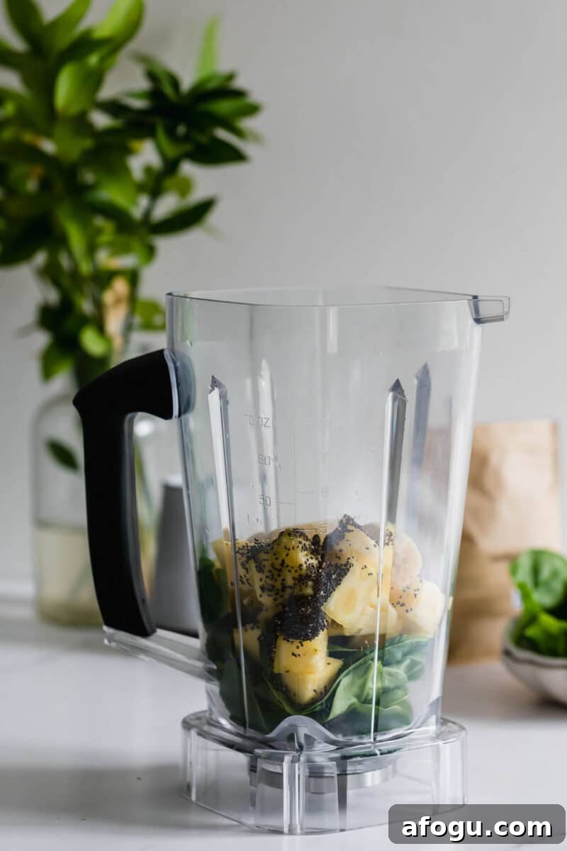 Dairy-Free Green Breakfast Smoothie Recipe 7 A blender pitcher containing spinach, pineapple chunks, banana pieces, and chia seeds sits on a white countertop.